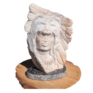 May include: A white marble sculpture of a Native American man wearing a headdress. The sculpture is detailed with feathers and a beaded necklace. The sculpture is sitting on a wooden base.
