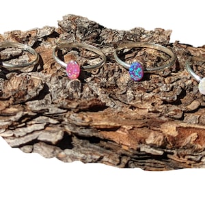 May include: Four silver rings with oval-shaped opal stones. The rings are arranged on a piece of tree bark. The opals are blue, pink, purple, and white.