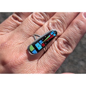 May include: A silver ring with a rectangular black inlay featuring a colorful geometric design. The inlay includes green, blue, red, and white shapes. The ring is shown on a hand.