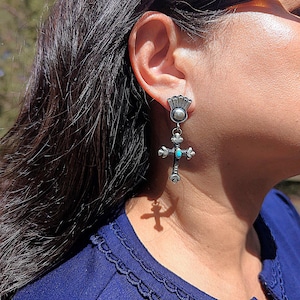 May include: A silver-toned cross earring with a small turquoise stone in the center. The earring has a decorative top piece and is worn on the ear. The cross design is detailed with ornate flourishes. The earring is shown against a blue background.