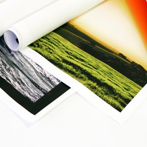 Op de afbeelding: Twee opgerolde afdrukken van landschapsfotografie. De ene afdruk toont een veld met groen gras met een zonsondergang op de achtergrond. De andere afdruk toont een zwart-witbeeld van een rivier.