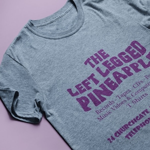 Puede incluir: Una camiseta gris jaspeada con texto morado que dice "THE LEFT LEGGED PINEAPPLE" e incluye el texto "Records-Tapes CD's-Po Music Vidoes & Compute T Shirts 24 CHURCHGATE TELEPHONE"