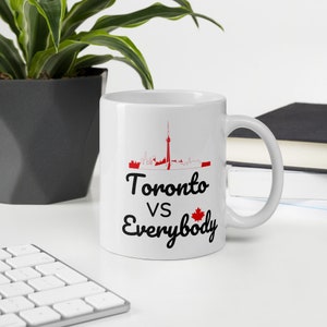 Puede incluir: Taza de cerámica blanca con un diseño en negro y rojo. El diseño presenta un horizonte de Toronto, Canadá, con la Torre CN y el texto "Toronto vs Everybody" con una hoja de arce canadiense.