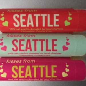 May include: Three lip balm tubes with a red, mint green, and pink background. Each tube has the text "kisses from SEATTLE" and "10% net profits donated to local charities. Collectible. Cruelty Free."