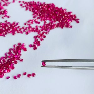 Puede incluir: Una colección de pequeñas gemas rojas vibrantes y redondas dispersas sobre una superficie blanca. Una gema es sostenida por unas pinzas plateadas. Las gemas son de un color rojo intenso y rico, y parecen pulidas.