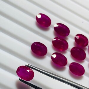 May include: A collection of oval-shaped, deep red gemstones, likely rubies, displayed on a white surface. One ruby is held by tweezers.