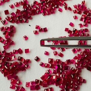 Puede incluir: Una imagen de primer plano de una gran cantidad de piedras preciosas sueltas, facetadas, de forma cuadrada, de color rojo rubí. Las piedras preciosas están esparcidas sobre una superficie blanca. Unas pinzas sujetan una de las piedras preciosas.
