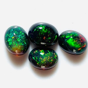 May include: Four oval-shaped black opals with green and red iridescent flashes. The opals are arranged in a cluster on a white surface.