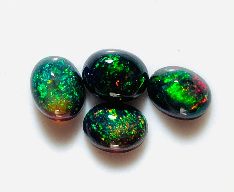 May include: Four oval-shaped black opals with green and red iridescent flashes. The opals are arranged in a cluster on a white surface.
