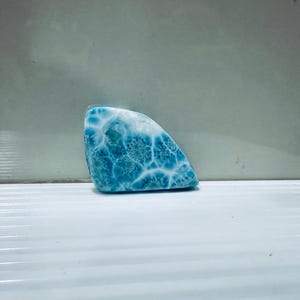 Puede incluir: Una gema pulida de forma triangular con un llamativo patrón azul y blanco. La superficie de la piedra muestra una red de intrincados diseños en forma de telaraña, creando una textura visual única. La paleta de colores está dominada por tonos azules y blancos.