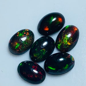 May include: Six oval-shaped black opals with green and red iridescent flashes. The opals are arranged in a cluster on a light gray surface.