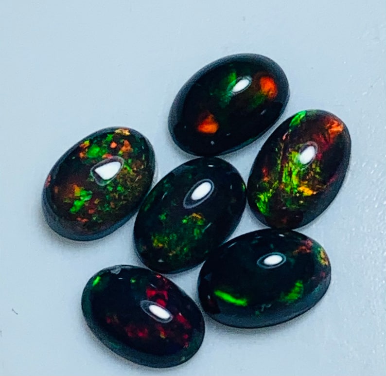 May include: Six oval-shaped black opals with green and red iridescent flashes. The opals are arranged in a cluster on a light gray surface.