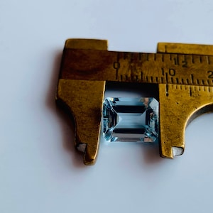 May include: A light blue gemstone, possibly aquamarine, is being measured with a brass caliper. The gemstone is rectangular in shape with slightly rounded edges. The caliper reads 10, 12, 20.