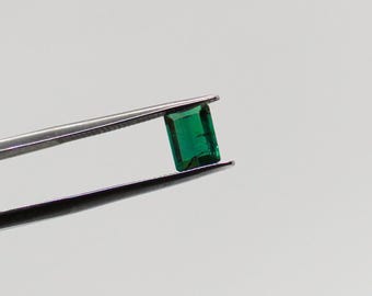 May include: A vibrant green emerald gemstone, cut into a rectangular shape, is held by tweezers against a white background.