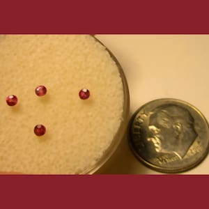 May include: Four small, round, red gemstones on a white, textured surface. A US dime coin is in the background for scale.