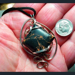 May include: A silver pendant with a black and gold marbled stone, wrapped in silver wire, hanging from a black cord. The pendant is displayed on a hand, next to a US dime.