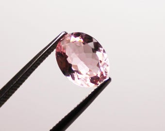 May include: A faceted, oval-shaped, light pink gemstone is held by a pair of black tweezers. The gemstone has multiple facets that reflect light, creating a sparkling effect. The background is a plain white.