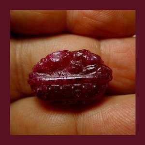 Puede incluir: Una piedra preciosa de rubí tallada de color rojo oscuro con forma de capullo de flor. La piedra preciosa se sostiene en una mano.