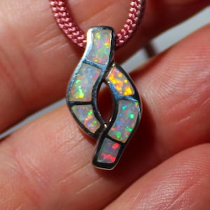 May include: A silver pendant with an abstract design, featuring iridescent opal inlays. The pendant hangs from a pink cord. The opal displays a range of colors, including green, yellow, and red, creating a vibrant and eye-catching piece of jewelry.