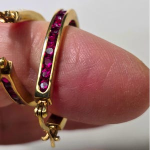May include: Close-up of gold hoop earrings with a row of small, vibrant red gemstones set along the front. The earrings are held up by a finger, showcasing the intricate design and the gleam of the gold.