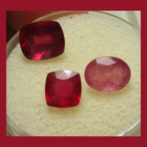 May include: Three red gemstones, two square and one oval, are displayed on a white surface.