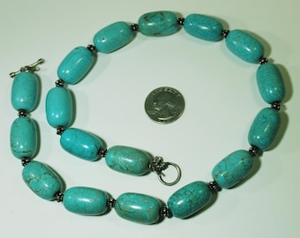 May include: A turquoise bead necklace with a silver clasp. The necklace features large, oval-shaped beads in varying shades of blue-green. The beads are strung together with small, dark metal beads. A coin is next to the necklace for size comparison.