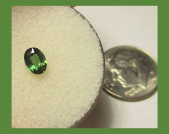 May include: A green oval gemstone on a white background, next to a US dime for size comparison.