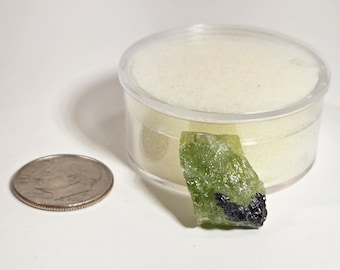 May include: A raw, green peridot gemstone with black inclusions, next to a dime and a small, clear plastic container. The peridot has a rough, natural texture and is a vibrant, translucent green color.