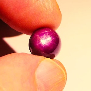 May include: A deep purple ruby gemstone with a star-like pattern, held between two fingers.