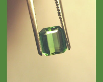 May include: A green emerald gemstone held by tweezers. The emerald is cut in a rectangular shape with beveled edges.