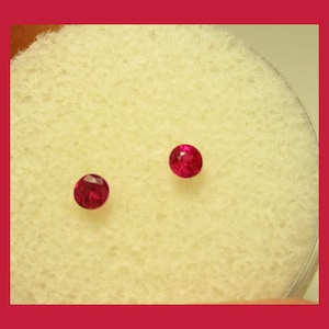 May include: Two round, red gemstones on a white, textured surface.