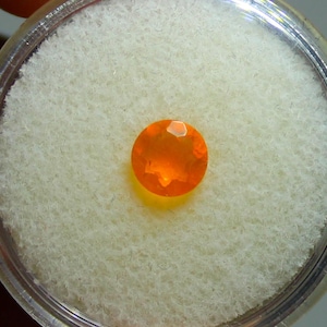 May include: A round, faceted, orange gemstone, possibly an opal, set against a white, textured background within a clear, circular container. The gemstone has a brilliant, fiery appearance.