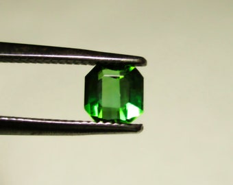May include: A vibrant green gemstone, cut into an octagonal shape, is held in tweezers. The gemstone is a deep, rich green color and has a smooth, polished surface.