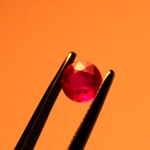 May include: A close-up of a red gemstone held by tweezers against an orange background.