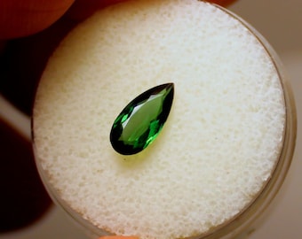 May include: A vibrant, pear-shaped, emerald-green gemstone. The gem is faceted, displaying a brilliant shine. It is set against a white, textured background within a clear container. The gemstone's color is a rich, deep green.