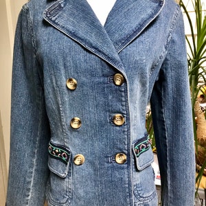 May include: A blue denim jacket with gold buttons and embroidered trim. The jacket has a double breasted closure and a pocket on the left side.