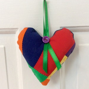 May include: A colourful patchwork heart-shaped ornament with a green ribbon and a purple button. The heart is made of different fabrics in shades of red, blue, green, orange, and yellow.