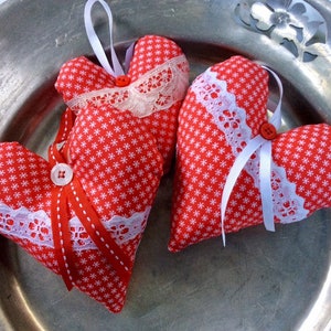 Puede incluir: Tres corazones de tela roja con patrones de copos de nieve blancos y ribete de encaje blanco. Cada corazón tiene una cinta blanca y un botón blanco.