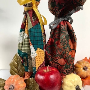 May include: Two fabric wine bottle gift bags with fall-themed patterns. One bag is a patchwork design with a yellow ribbon. The other bag has a leafy pattern with a black and white ribbon. The bags are surrounded by fall decor, including apples, pumpkins, and leaves.
