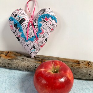 May include: A pink heart-shaped pillow with a white background and a blue and pink trim. The pillow is decorated with a pattern of cats and stars. The pillow is hanging from a pink ribbon and is sitting on a piece of driftwood next to a red apple.