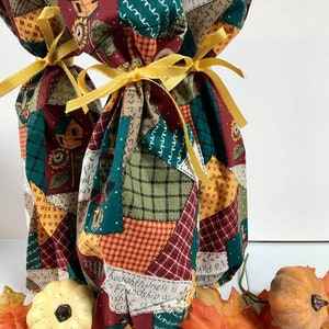 Può includere: Due sacchetti regalo per bottiglie di vino realizzati in tessuto patchwork in una gamma di colori verde, rosso e marrone. I sacchetti sono legati con un nastro giallo e presentano una varietà di disegni stampati, tra cui case, girasoli e testo. I sacchetti sono su una superficie bianca con foglie autunnali e piccole zucche.