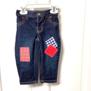 Könnte beinhalten: Dunkelblaue Jeans mit drei bunten Flicken. Die Flicken sind rot-weiß-kariert, rot und blau-weiß-kariert. Die Jeans hängen an einem weißen Kleiderbügel.