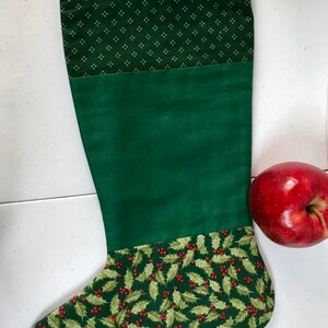 Può includere: Calza di Natale verde con la parte superiore a motivi bianchi e verdi, una sezione centrale verde unita e una parte inferiore verde con un motivo di foglie di agrifoglio e bacche rosse.