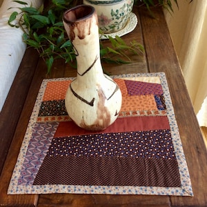 Può includere: Un runner da tavolo in patchwork marrone, bianco e arancione con un vaso in ceramica bianca con segni marroni in cima.