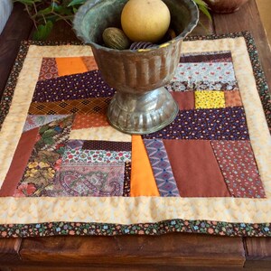 Può includere: Un runner da tavolo in patchwork con uno schema di colori marrone, arancione e giallo. Il runner è realizzato con vari tessuti con motivi floreali e paisley. Una ciotola di metallo con frutta è posizionata sopra il runner.