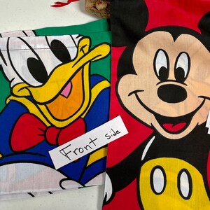 May include: Two fabric drawstring bags with cartoon characters. The bag on the left features Donald Duck in blue and yellow with a red bow tie. The bag on the right features Mickey Mouse in red, black, and yellow. Both bags have handwritten labels with the characters' names and measurements in inches.