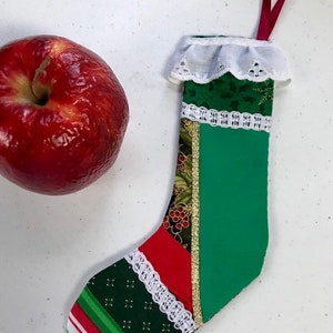 May include: A handmade Christmas stocking with a red ribbon loop. The stocking features green, red, and patterned fabric with white lace trim and a gold stripe. A red apple is next to the stocking.