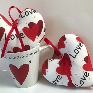 Peut inclure: Deux coussins en forme de cœur en tissu blanc avec des cœurs rouges et le mot "Love" imprimé dessus. Les coussins sont posés sur une tasse blanche avec un motif de cœur rouge et de flèche.