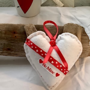 May include: White felt heart-shaped cushion with red ribbon and white polka dots. The ribbon has a red button and the text "Be Mine" on it.