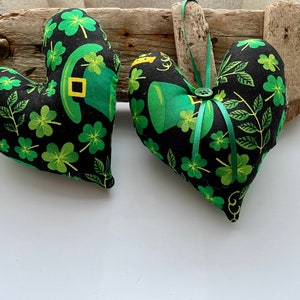 May include: Two black fabric heart-shaped cushions with a green shamrock and leaf pattern. The cushions have a green felt shamrock and a gold buckle on them.